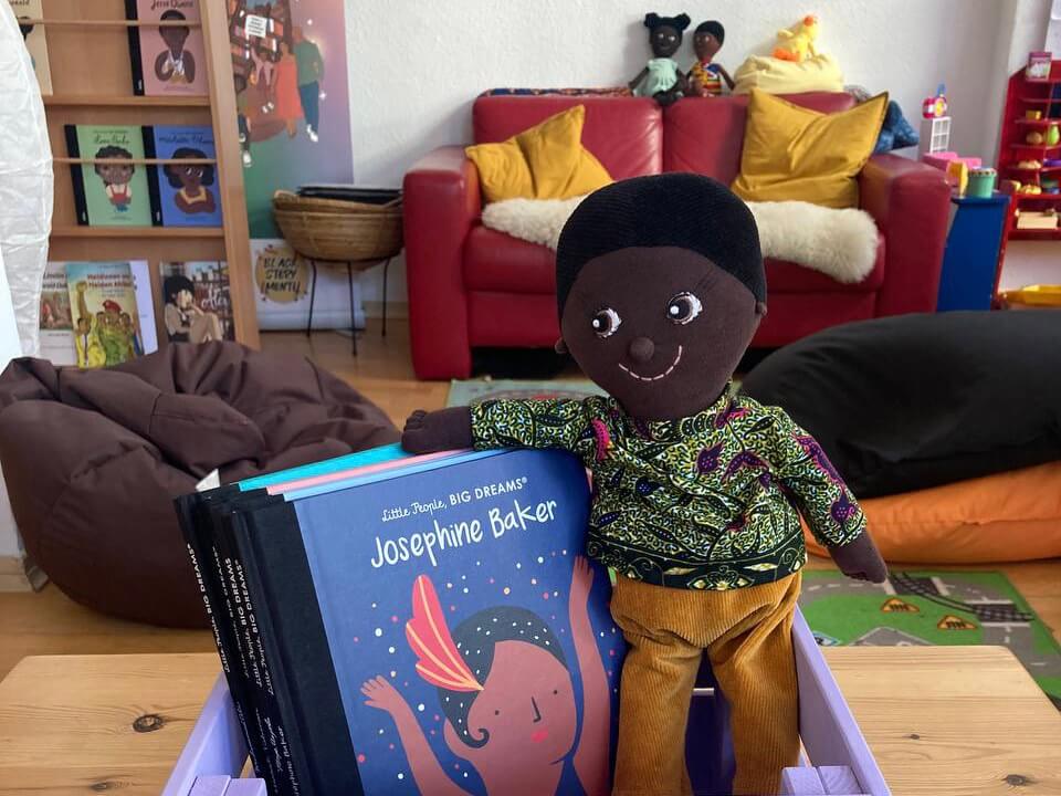 Stoffpuppe neben Kinderbuch „Josephine Baker“ in einem farbenfrohen Kinderzimmer mit Büchern im Hintergrund.